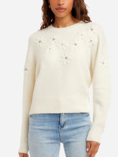 Woman wearing a cream sweater with floral embellishments and light blue jeans on a white background
