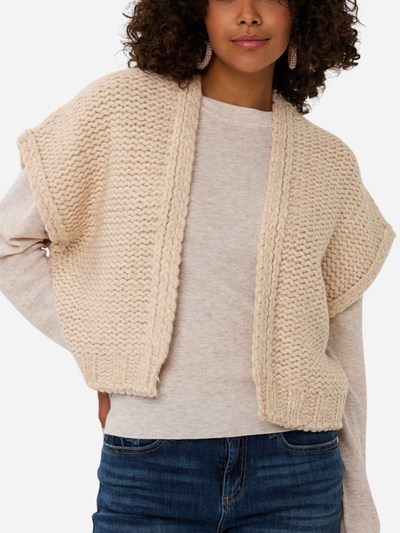 Woman wearing a beige knitted vest over a light gray shirt and blue jeans on a white background